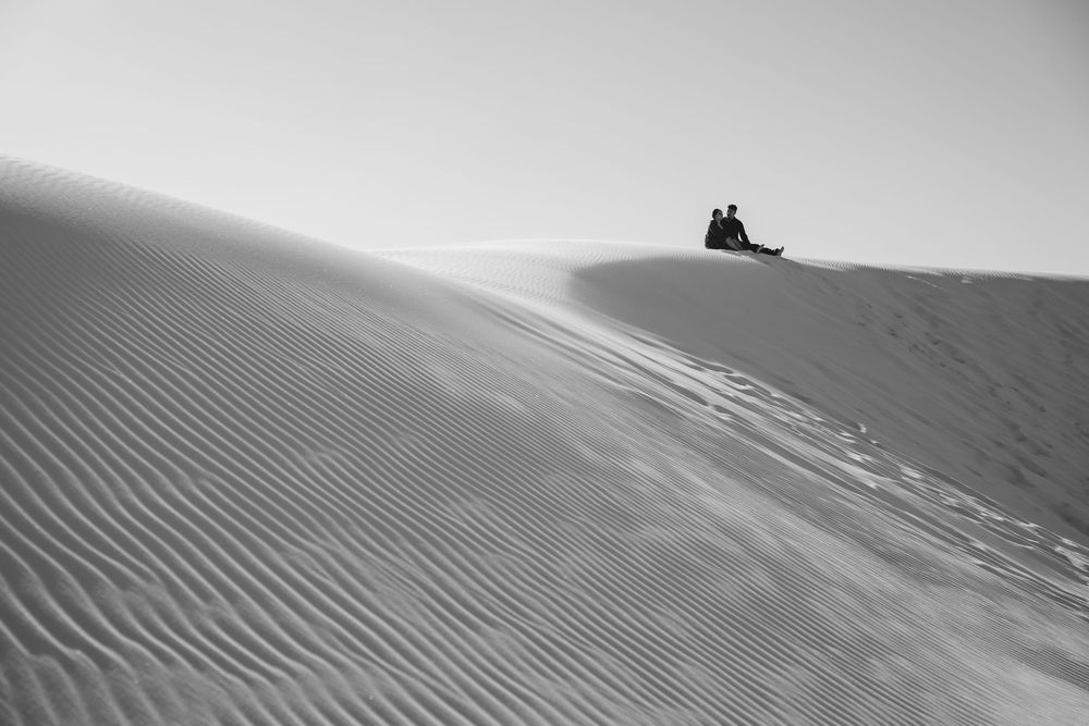 Two people sit atop a vast sand dune in a striking black and white desert landscape photo.