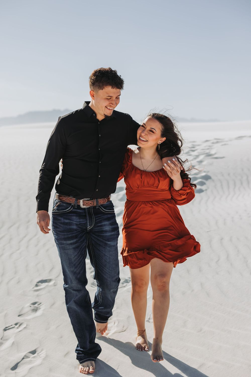 A happy couple walks barefoot on white sand dunes, woman in red dress and man in black shirt and jeans embracing.