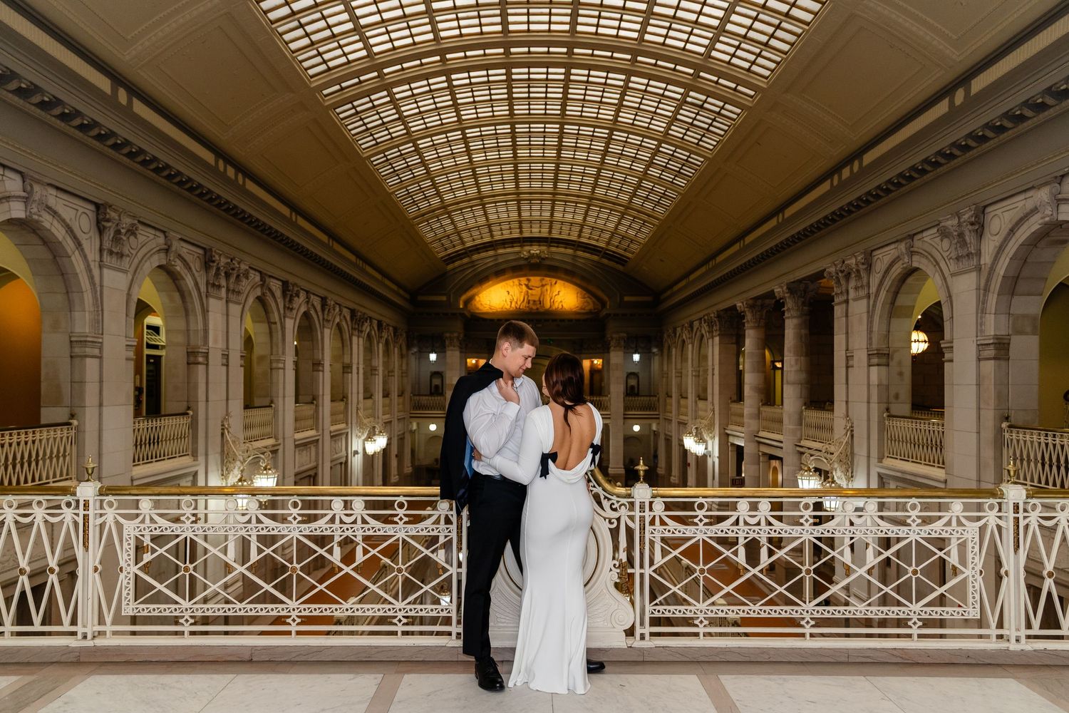 Elegant couple in wedding attire holding hands while ascending ornate marble staircase in grand historic building.