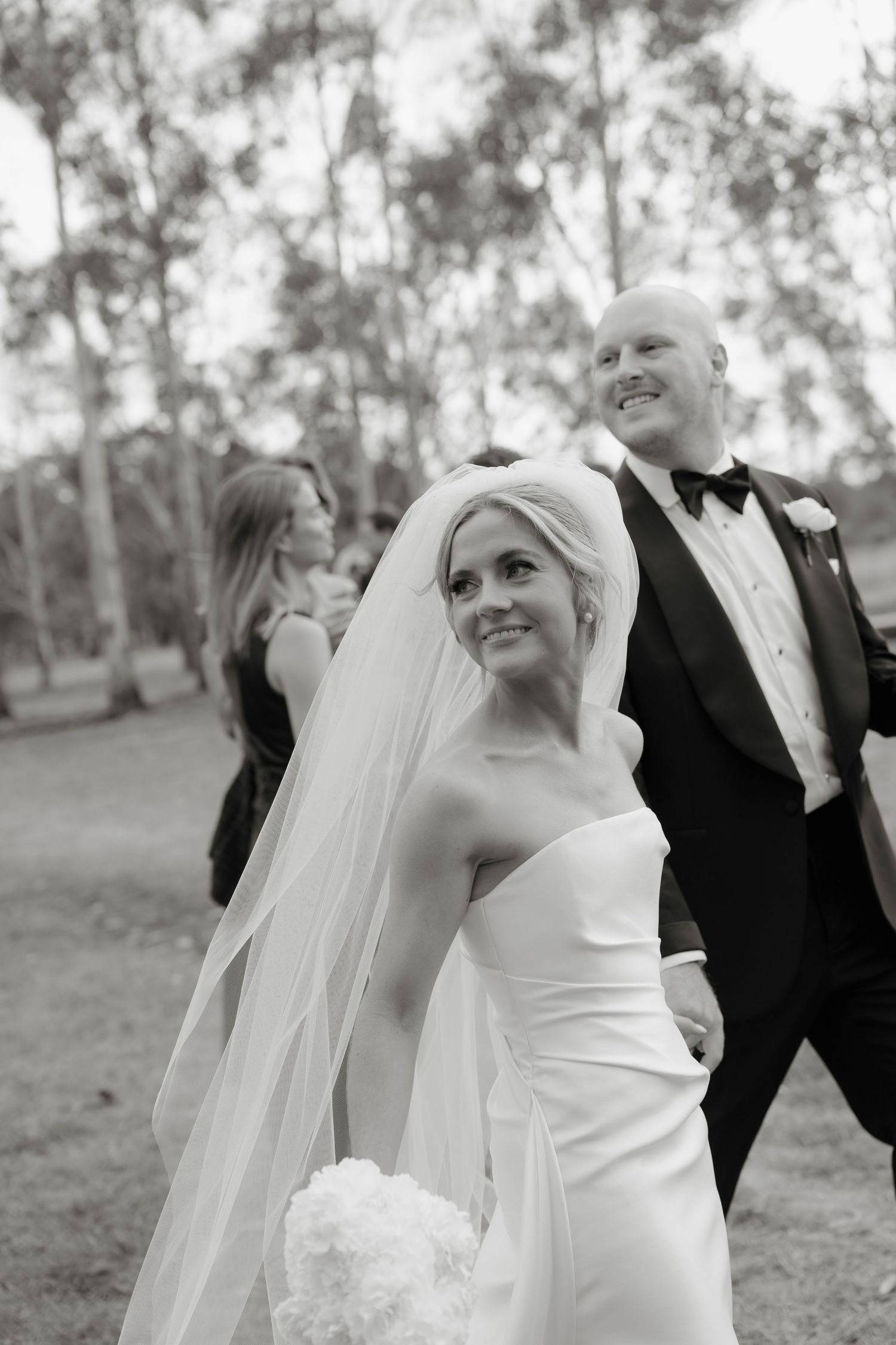 Bride in white gown and groom in white jacket walk under clear umbrella in lush green orchard on rainy wedding day.