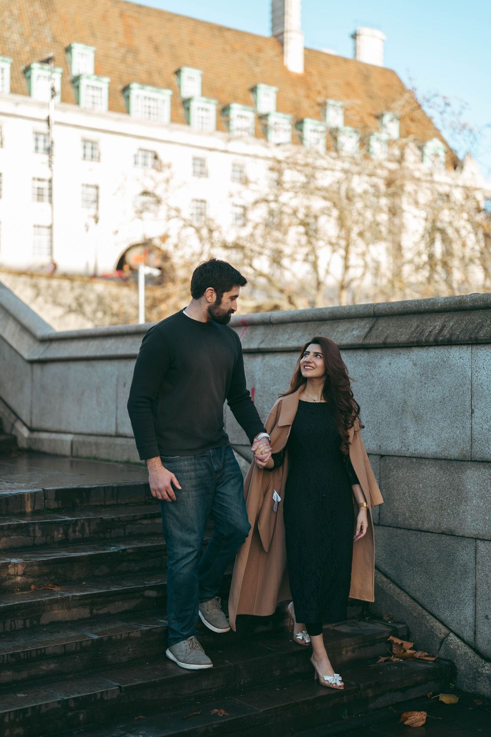 Couple holding hands walking down stone steps near historic building in autumn.