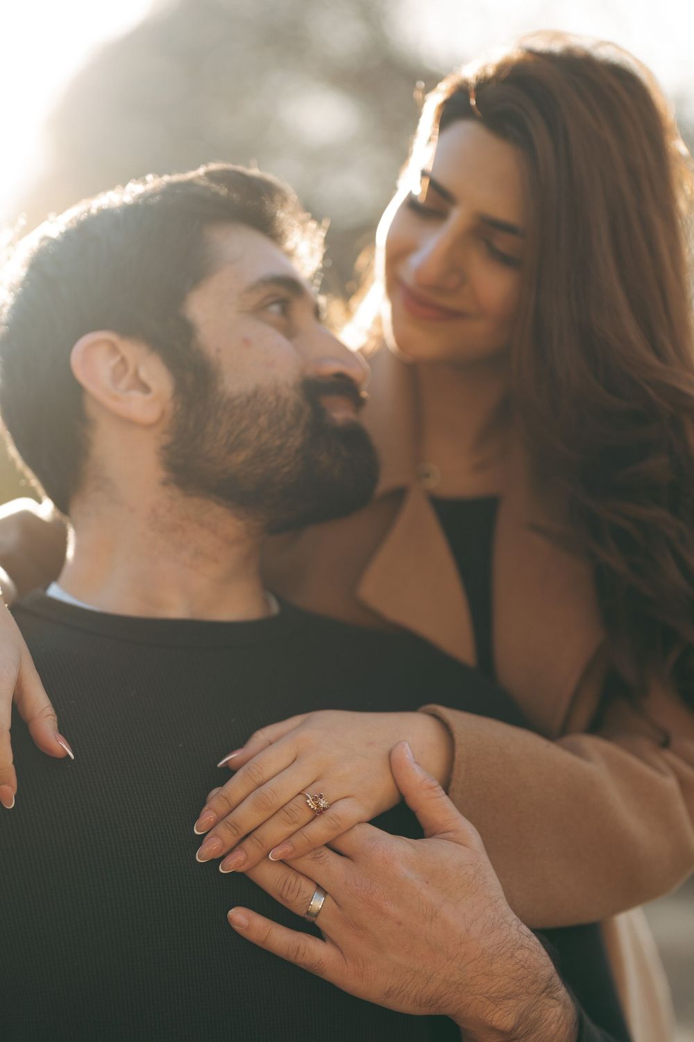 Couple sharing a romantic moment outdoors, woman wearing engagement ring, bathed in warm golden sunlight.