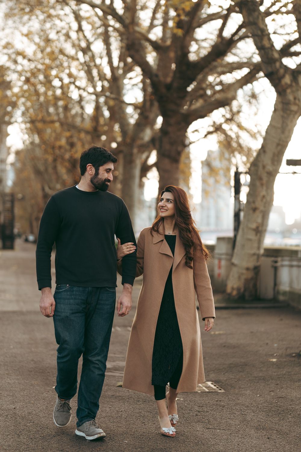 Couple walking together in autumn park, woman in beige coat smiling at bearded man in black sweater and jeans.