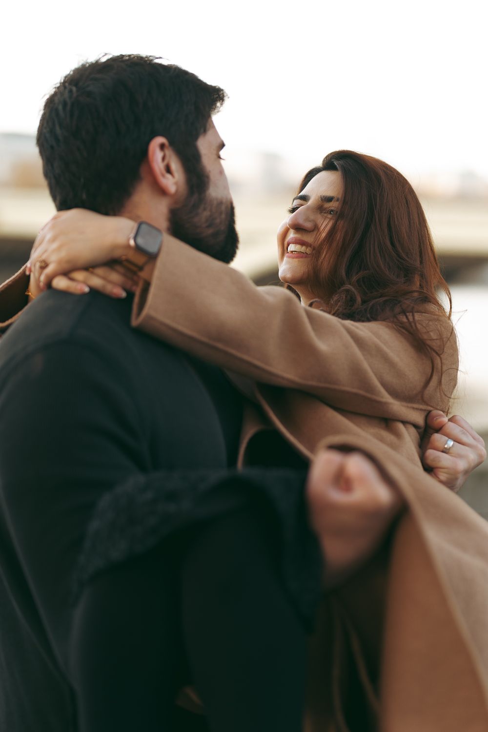 A joyful woman in a beige coat embraces a bearded man outdoors, laughing and looking into his eyes.