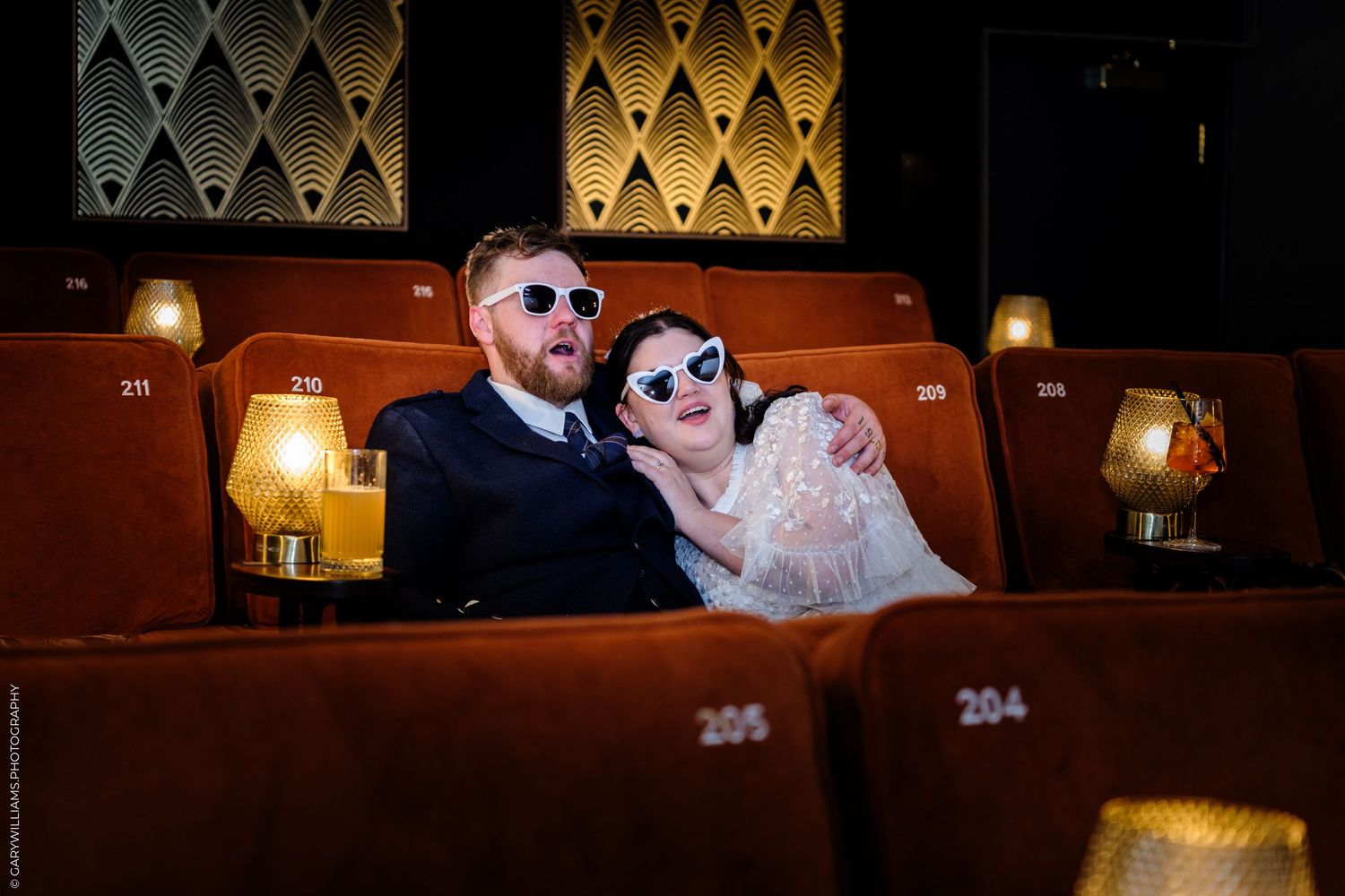 Bride and groom wearing heart sunglasses sitting in vintage red cinema seats in Cinema at The Hub St Albans