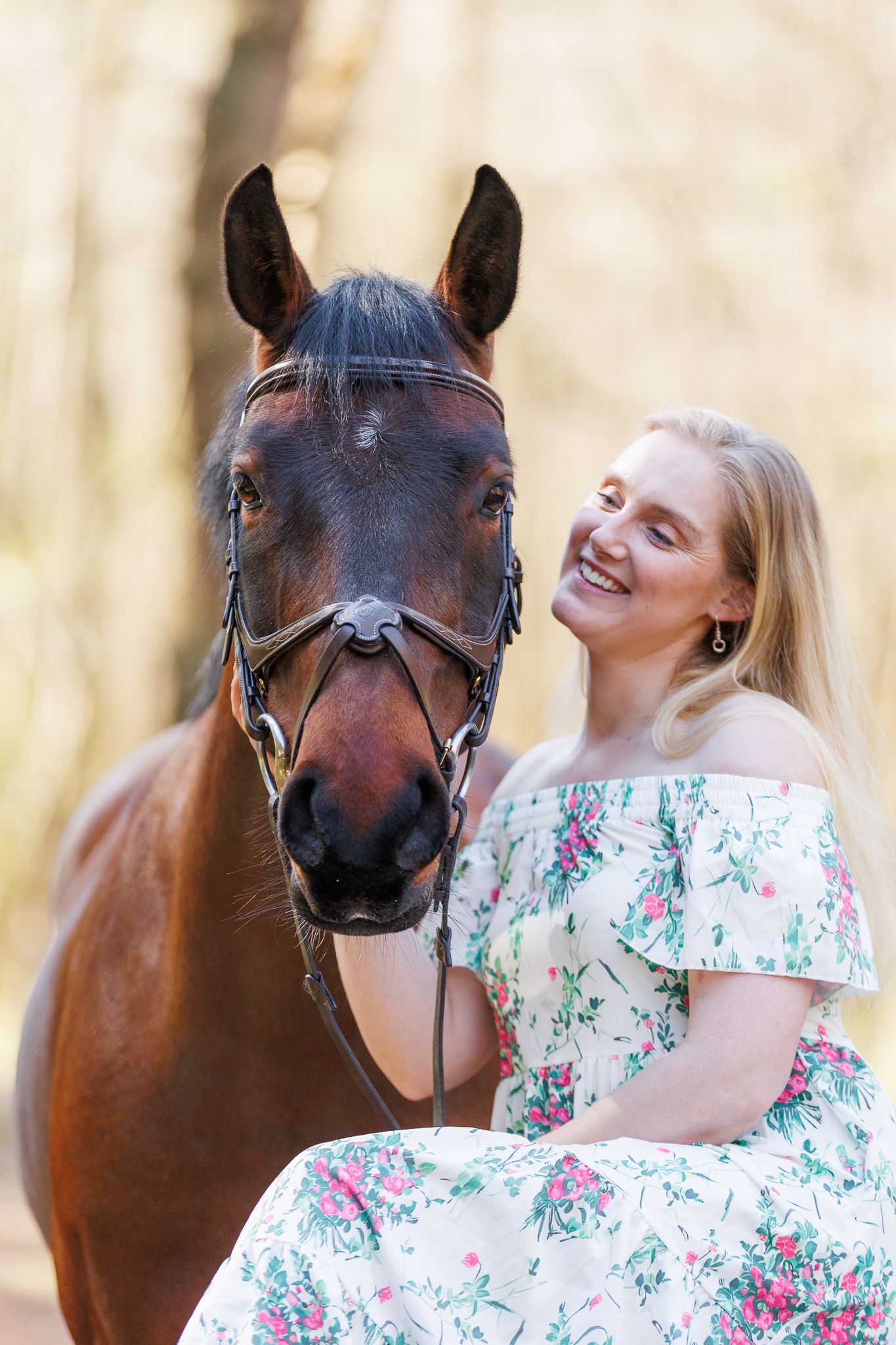 Smiling blonde woman in floral dress posing with a brown horse in a sunlit forest setting.