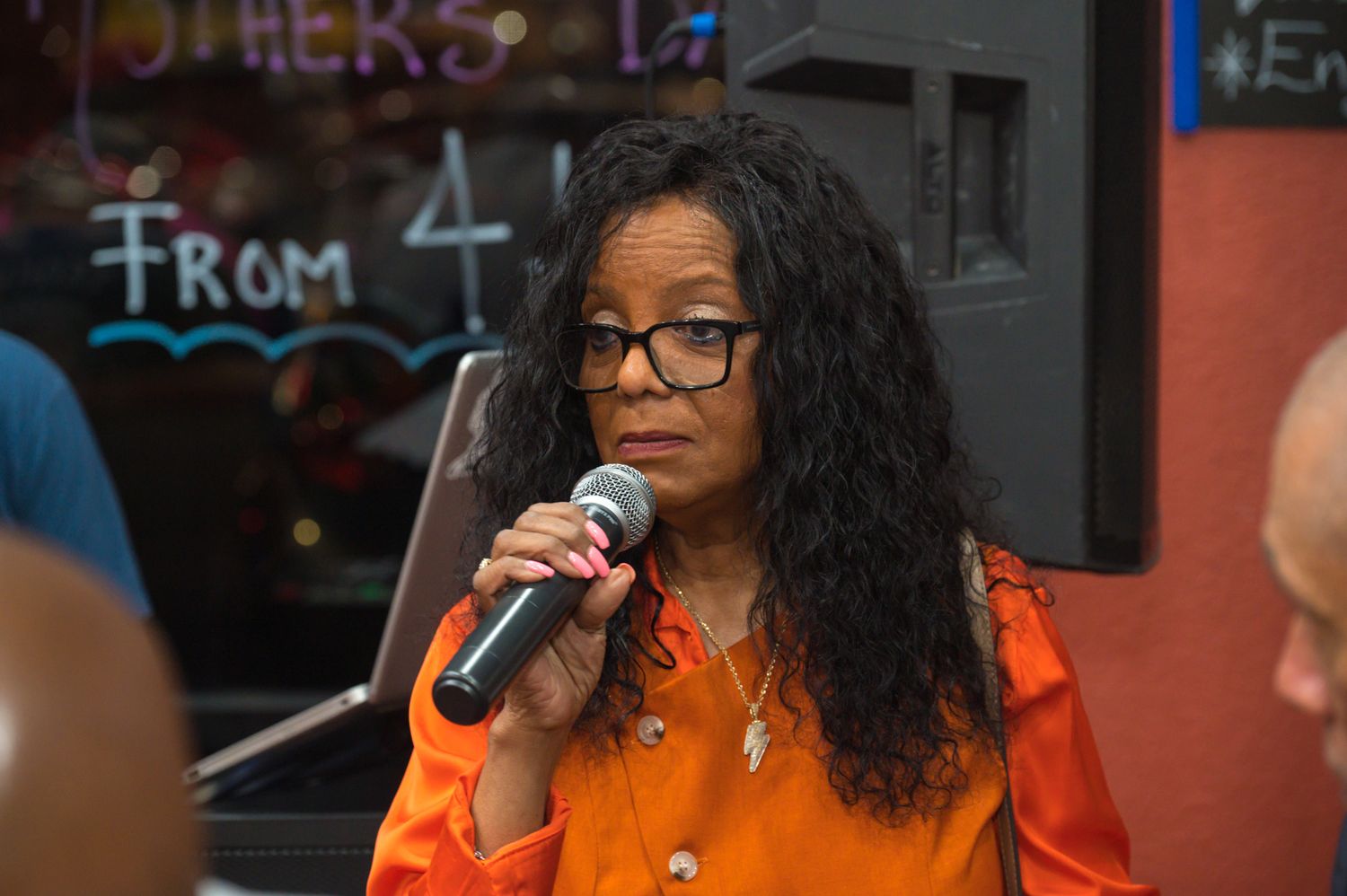 Woman in orange outfit speaking into microphone at an indoor event with colorful chalkboard background.