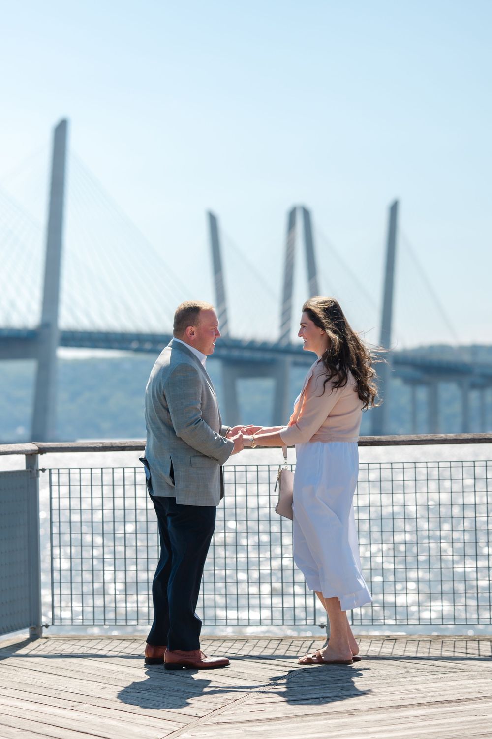 Couple holding hands on a waterfront deck with a cable-stayed bridge backdrop on a sunny day.