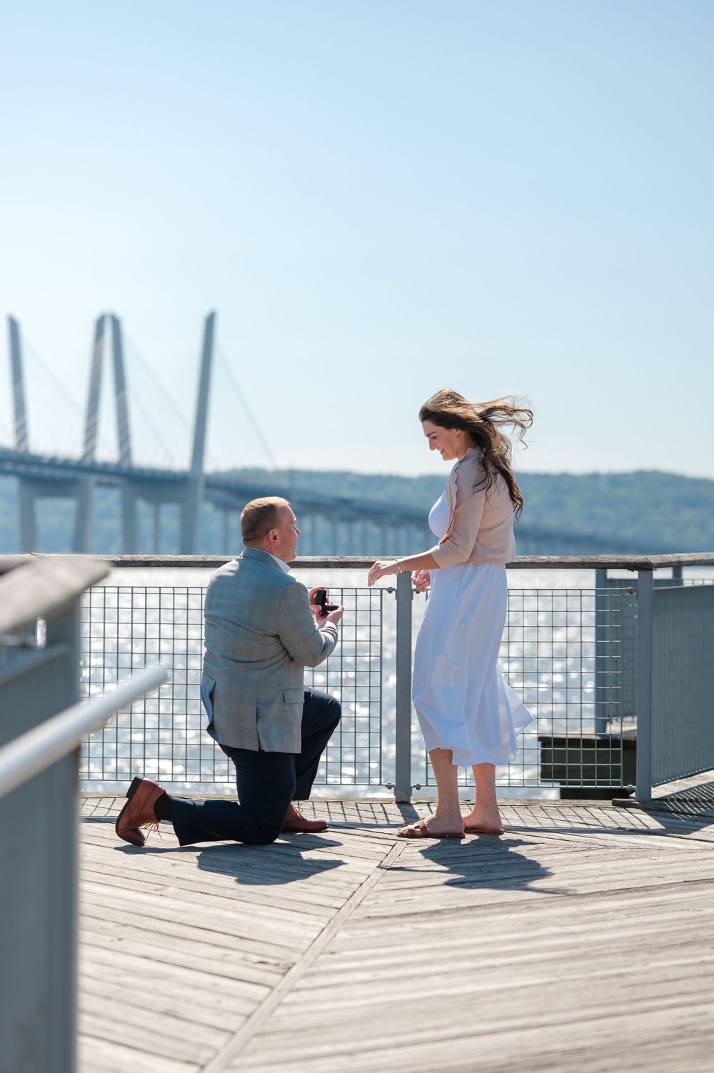 Man proposing on one knee to woman on a waterfront deck with a bridge in the background on a sunny day.