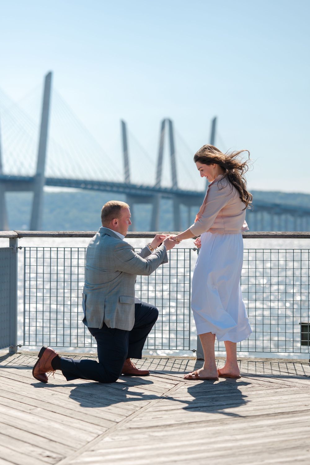 Man proposing on one knee to woman on a waterfront deck with a cable-stayed bridge in the background.