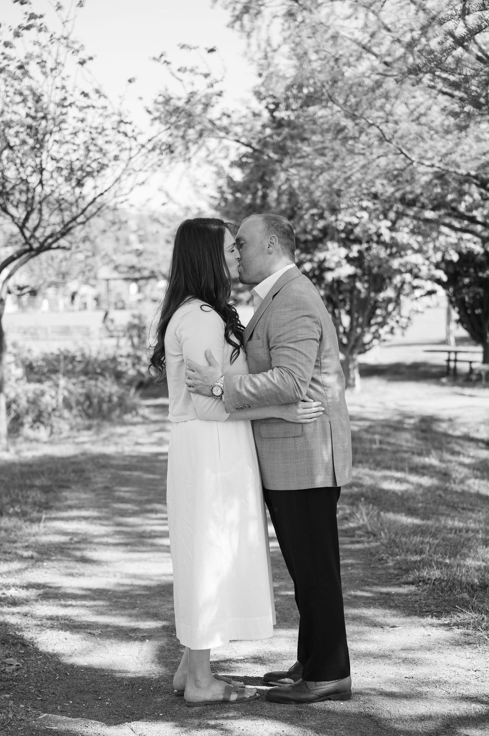 Black and white photo of a couple sharing a kiss outdoors among blooming trees, woman in white dress, man in suit jacket.
