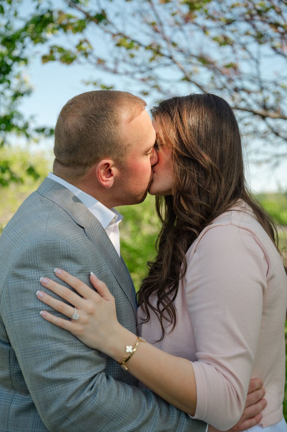 A couple shares a romantic kiss outdoors during an engagement photo session, with green trees in the background.