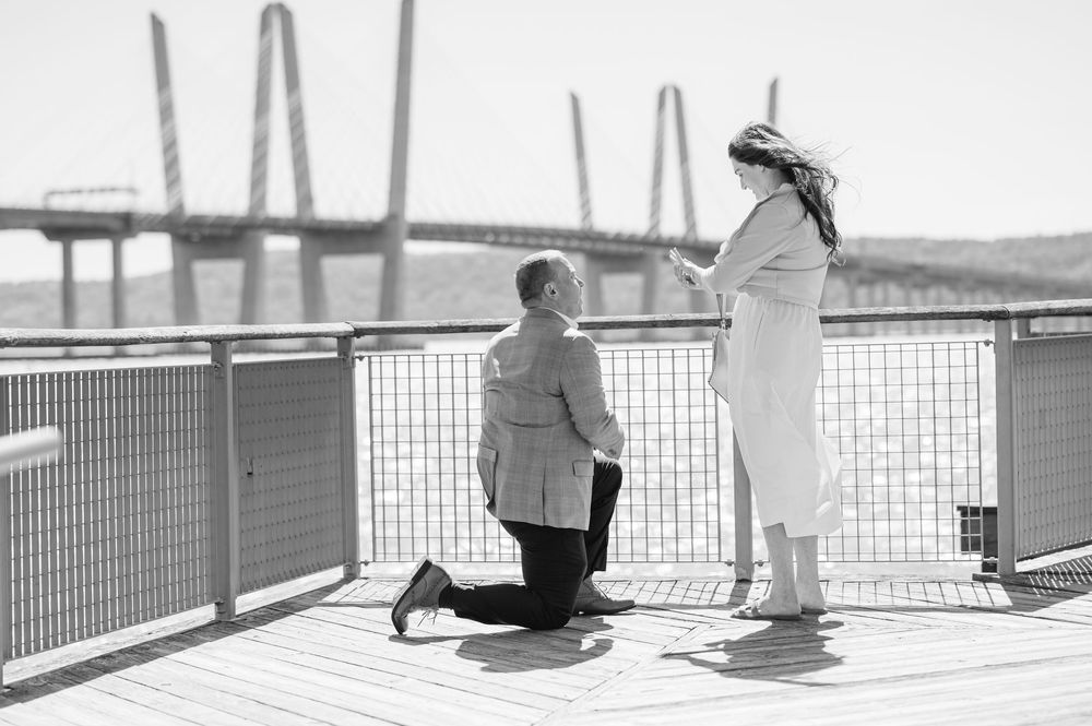 Man proposes on one knee to woman on waterfront deck with iconic cable-stayed bridge in black and white photo.