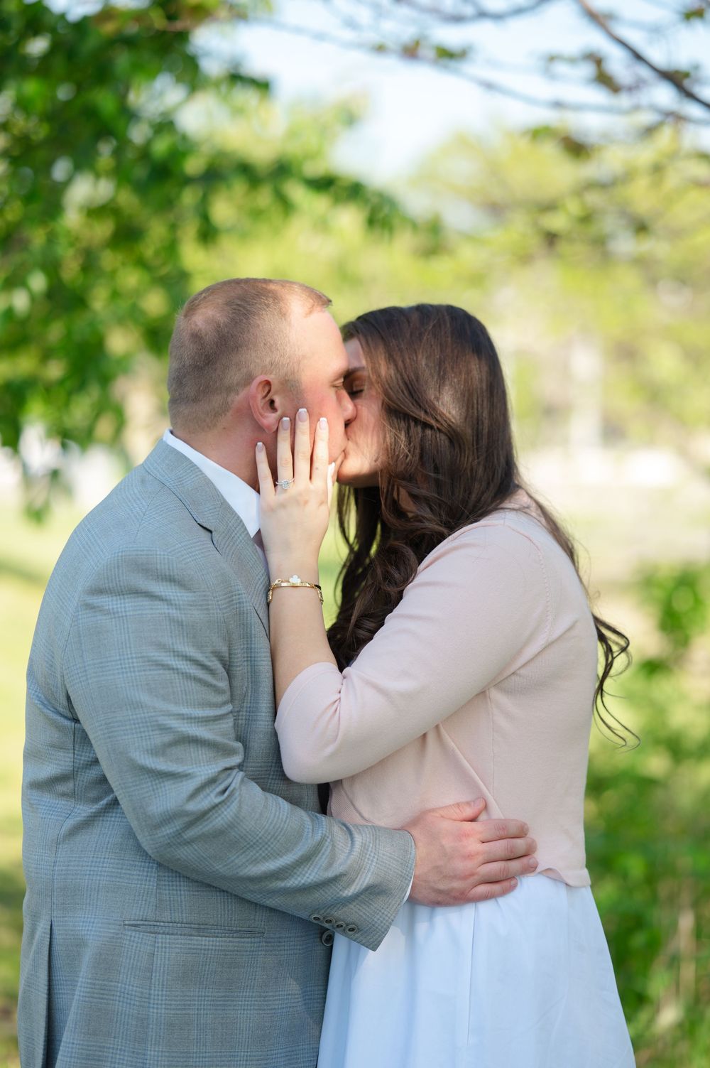Couple sharing a romantic kiss outdoors, woman in pink blazer and white skirt, man in gray suit, green trees background.
