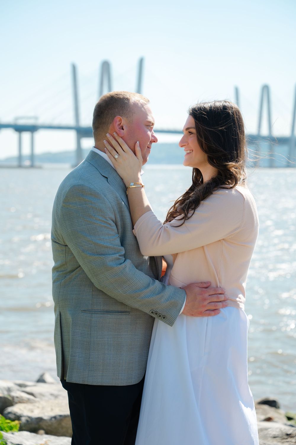 Couple embracing romantically by the waterfront with a cable-stayed bridge in the background on a sunny day.