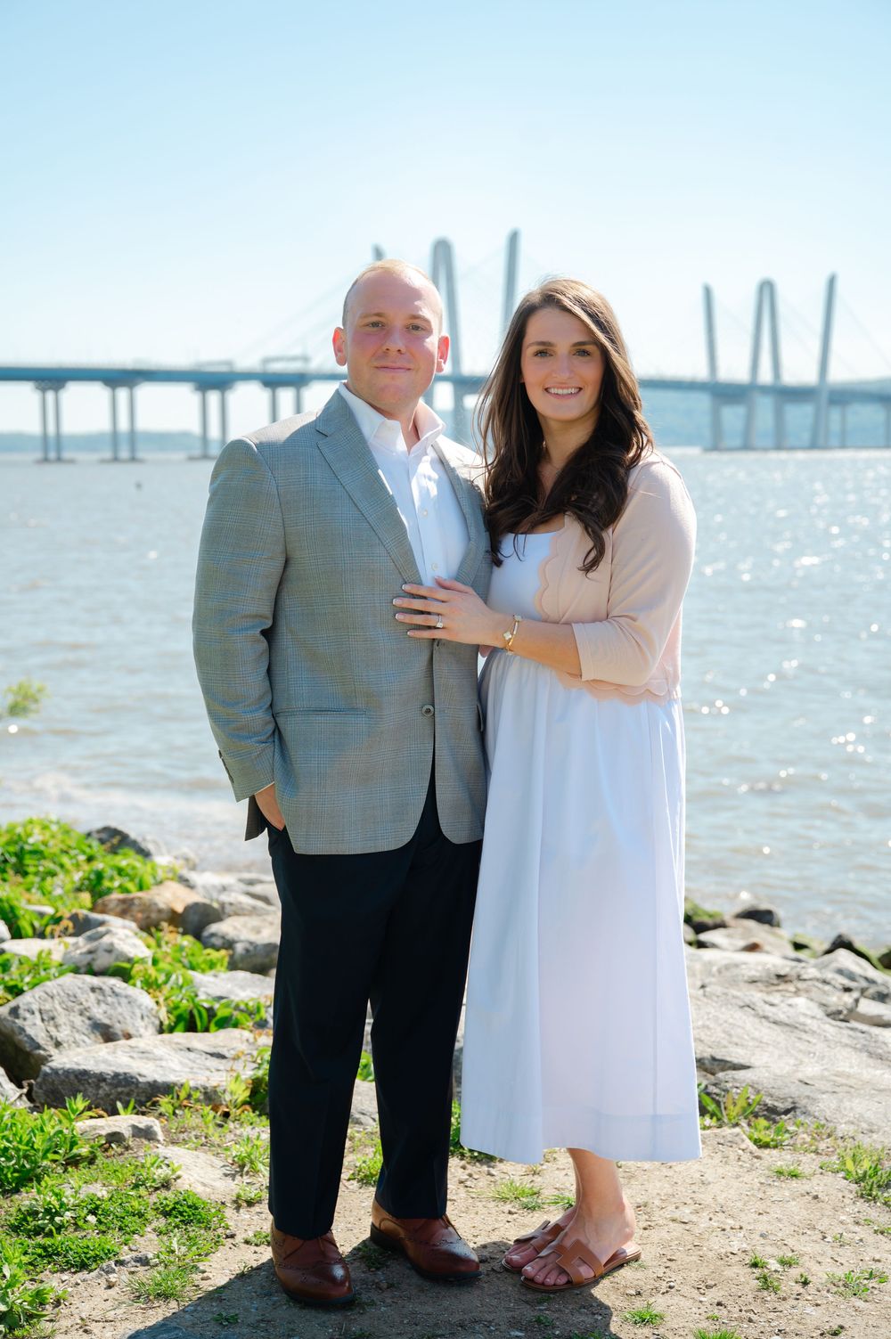 Couple posing by waterfront with bridge backdrop on sunny day, man in gray blazer, woman in white dress.