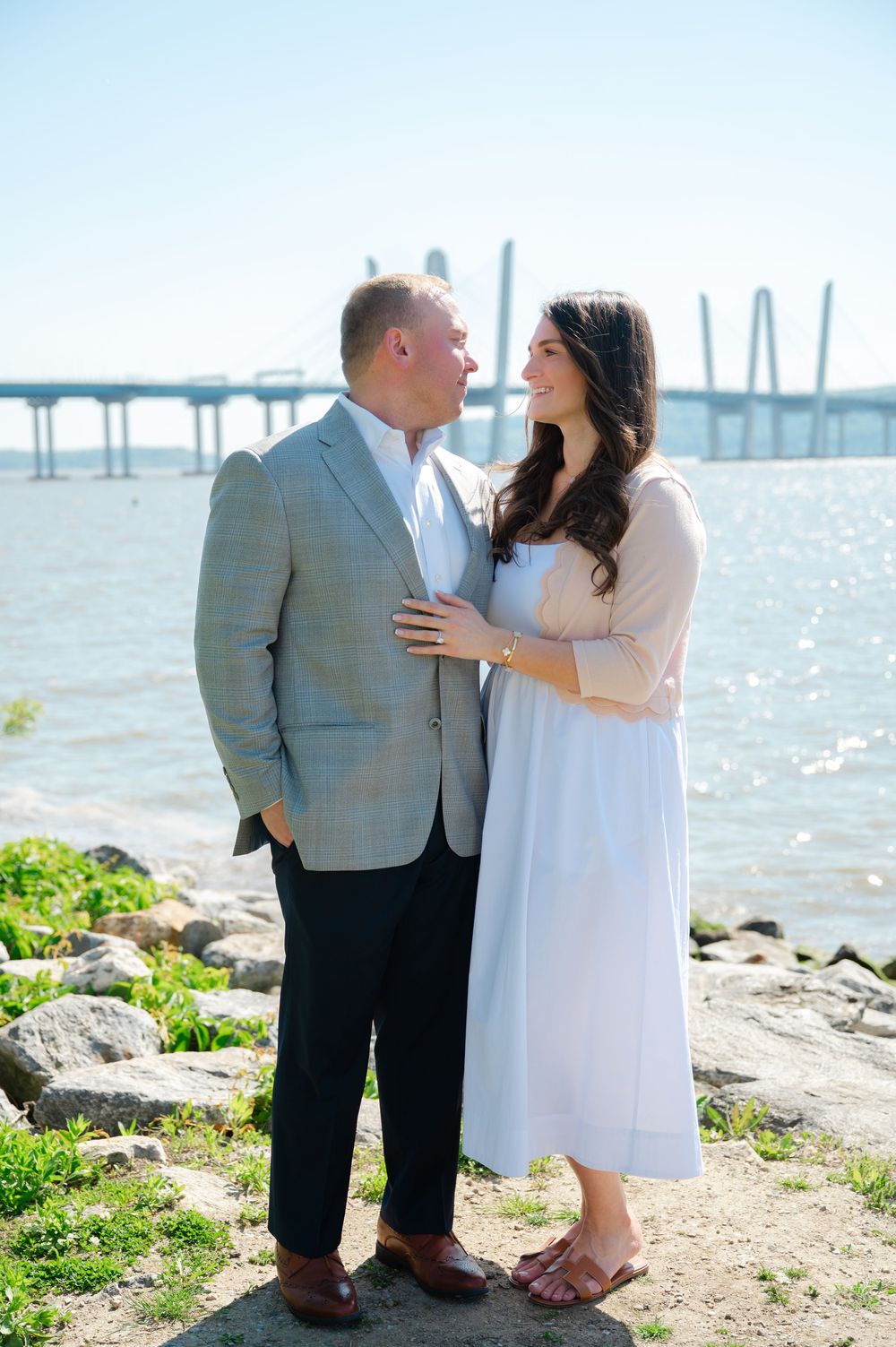 Couple sharing a romantic moment by the waterfront with a bridge in the background on a sunny day.