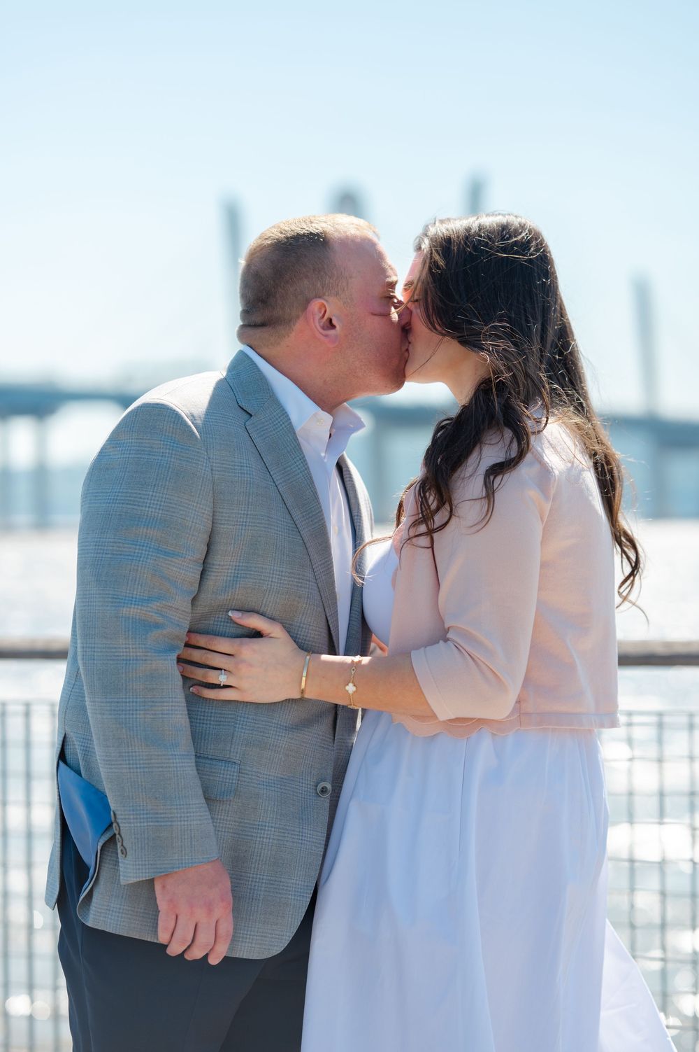Couple sharing a romantic kiss outdoors near a waterfront bridge on a sunny day, man in gray blazer and woman in white dress.