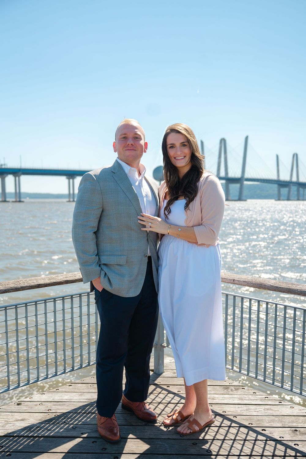Engaged couple posing on waterfront dock with suspension bridge behind them on a sunny day.