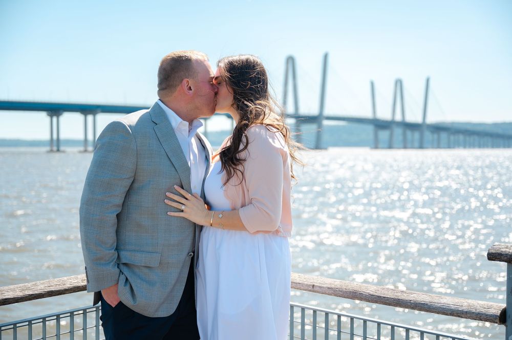 Couple sharing a romantic kiss by the waterfront with a cable-stayed bridge sparkling in the background.