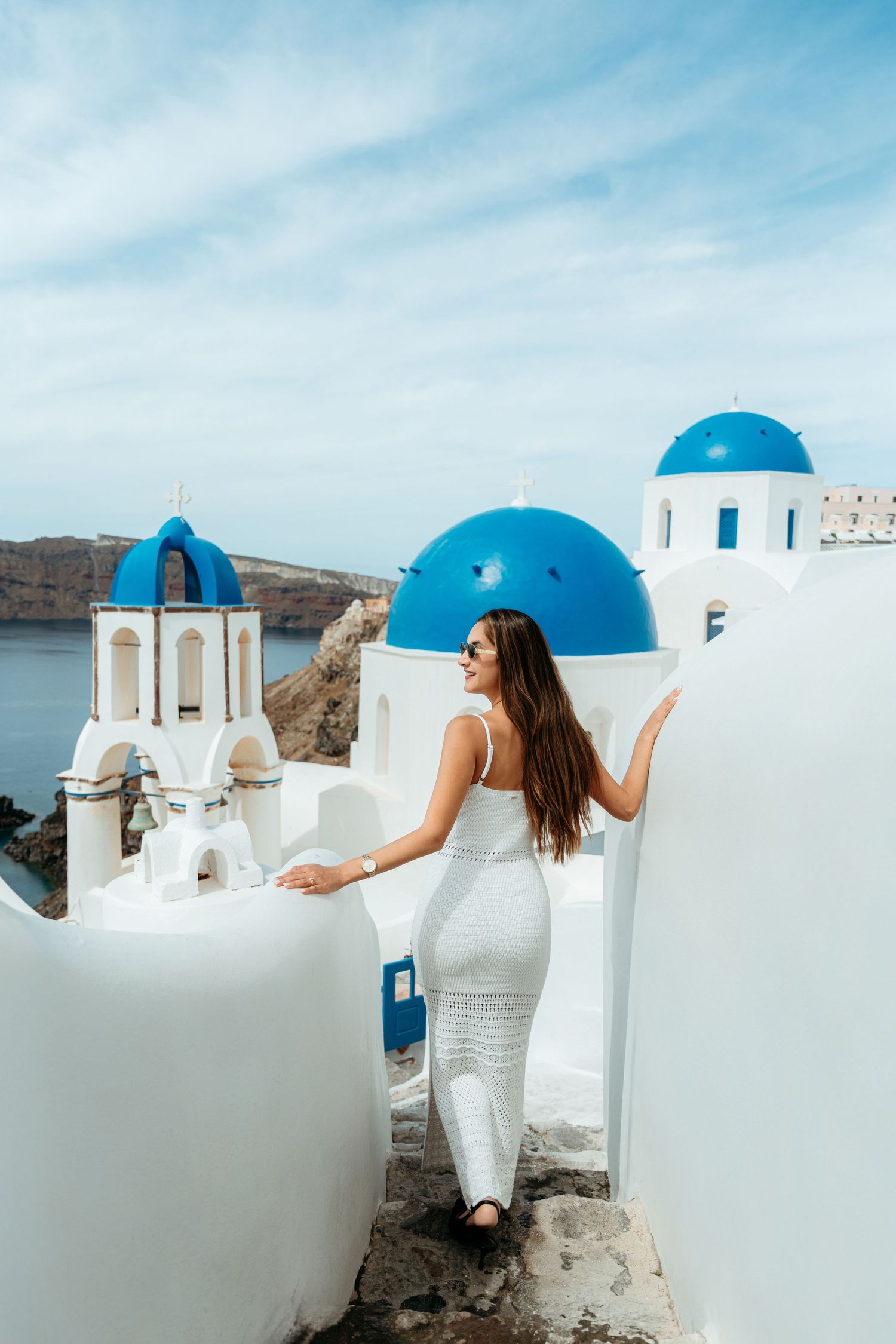 Woman in white dress walking among iconic blue-domed churches in Oia, Santorini, Greece, with caldera views.