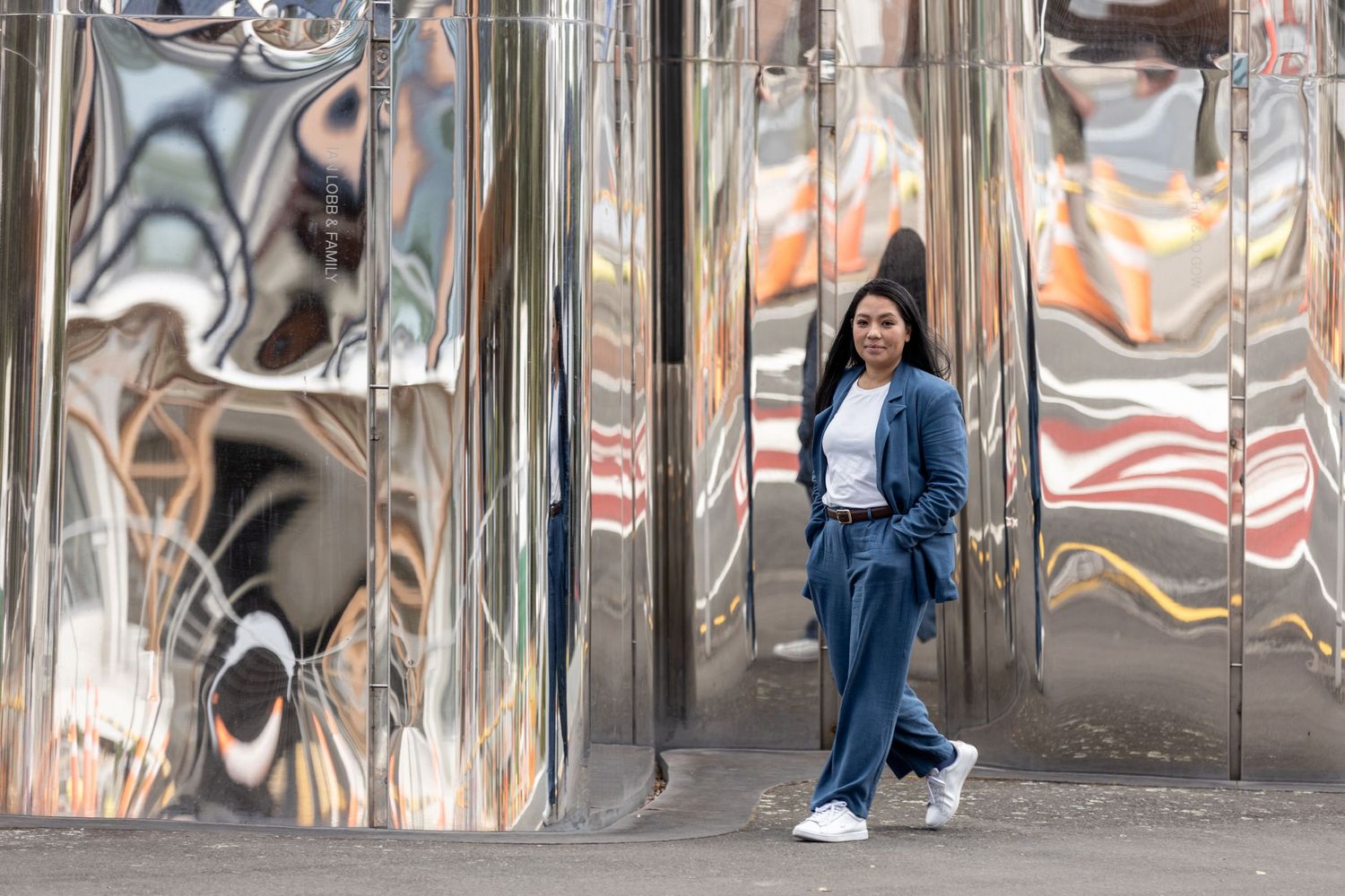 Woman in blue suit poses beside large reflective chrome sculptures on a city street.