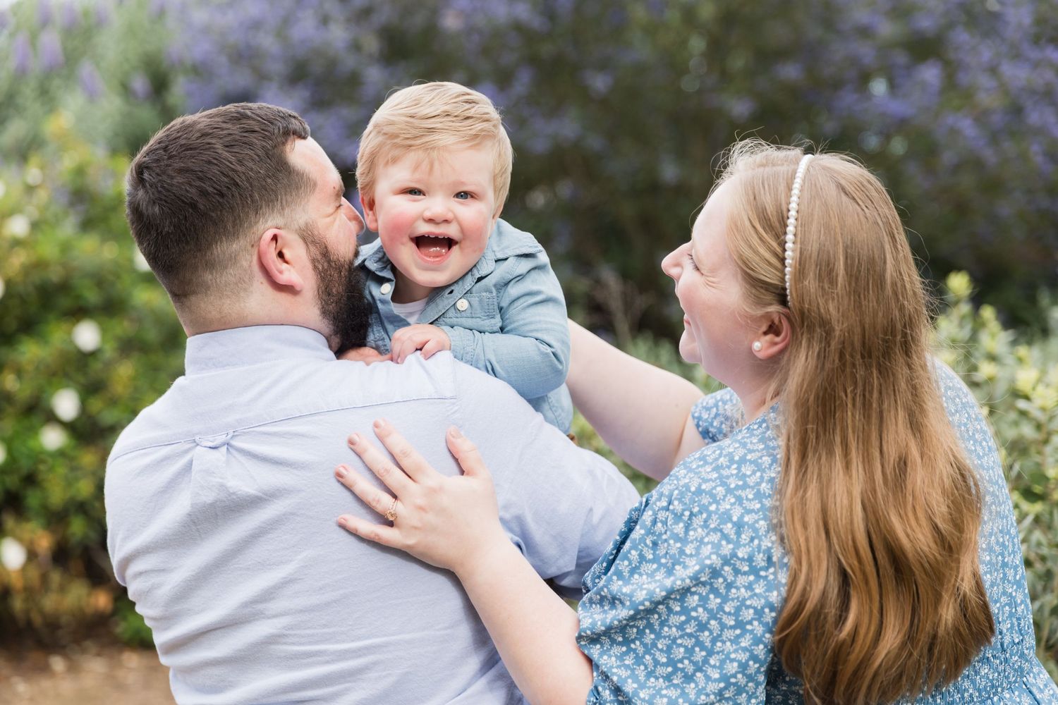 Happy baby laughing while being held by smiling parents outdoors near purple flowering bushes.