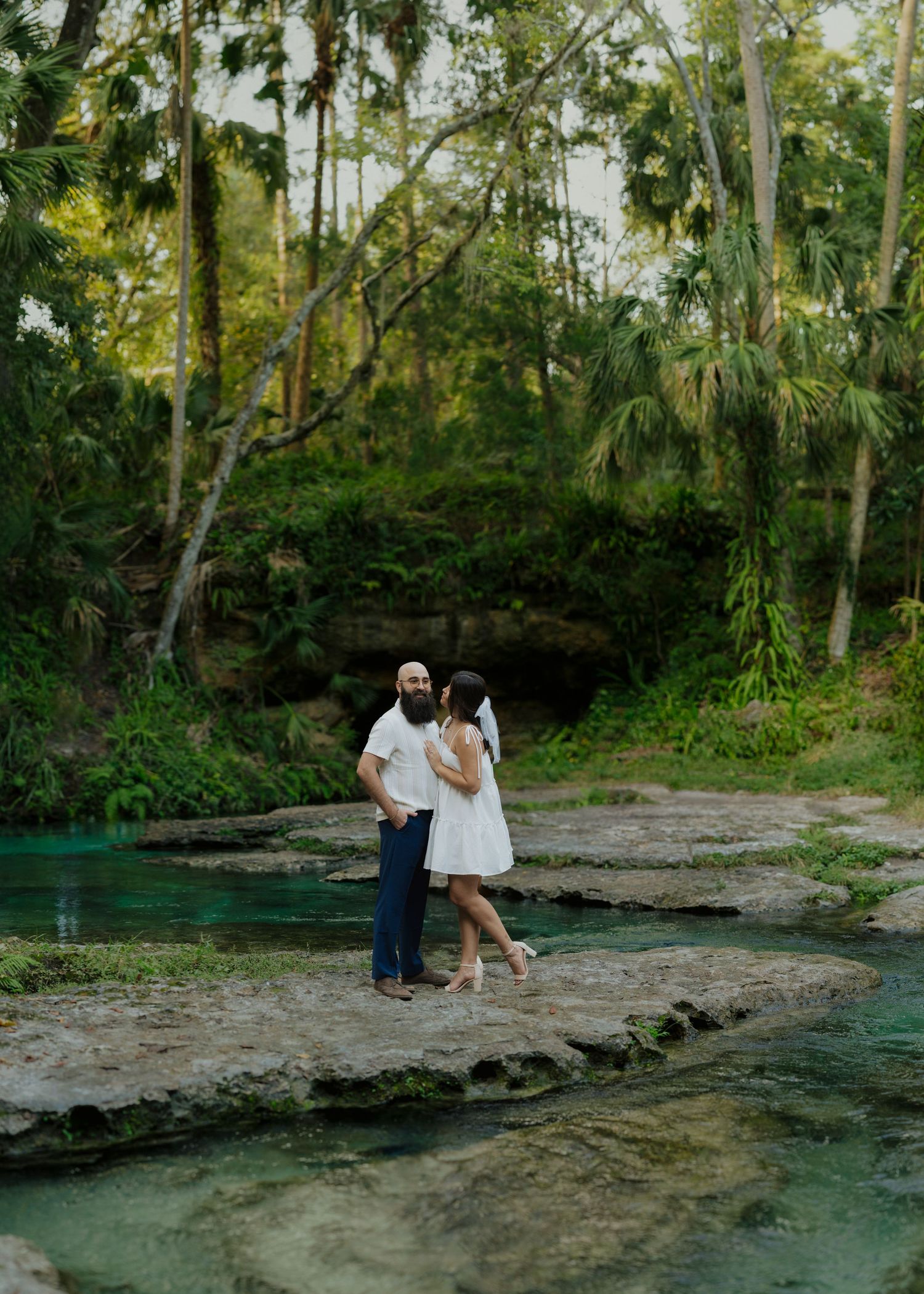 Couple standing on rocky shoreline beside turquoise spring water surrounded by lush tropical trees.