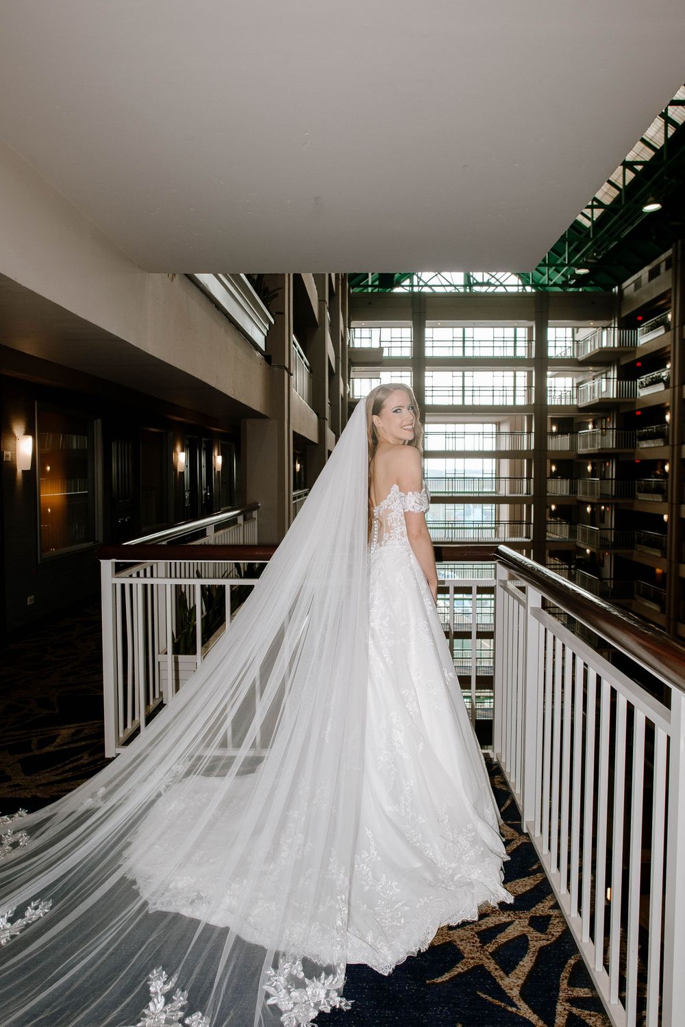 Bride in lace wedding gown with cathedral veil on hotel balcony overlooking atrium.