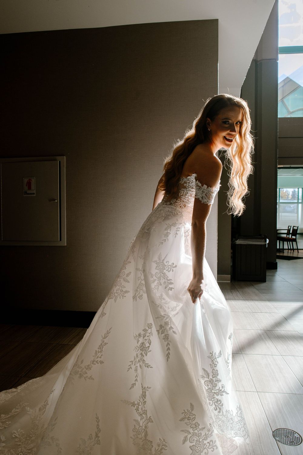 Bride in off-shoulder lace wedding dress smiling in dramatic natural light indoors.