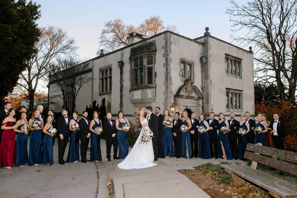 Large wedding party poses in navy and black formal attire outside a historic stone building.