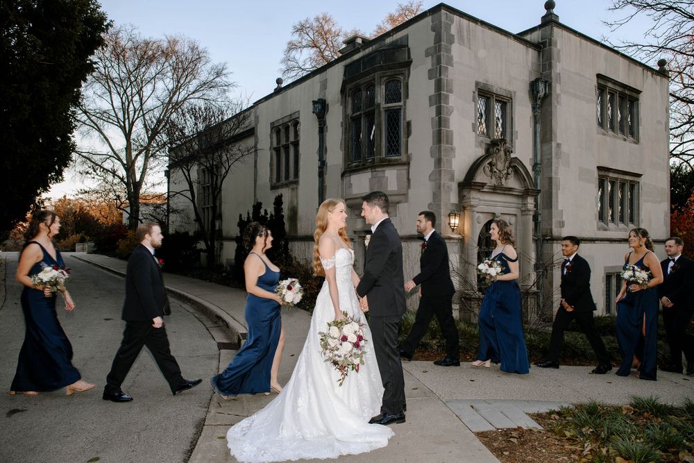 Bride in white gown walks with wedding party in navy dresses past a charming stone building.