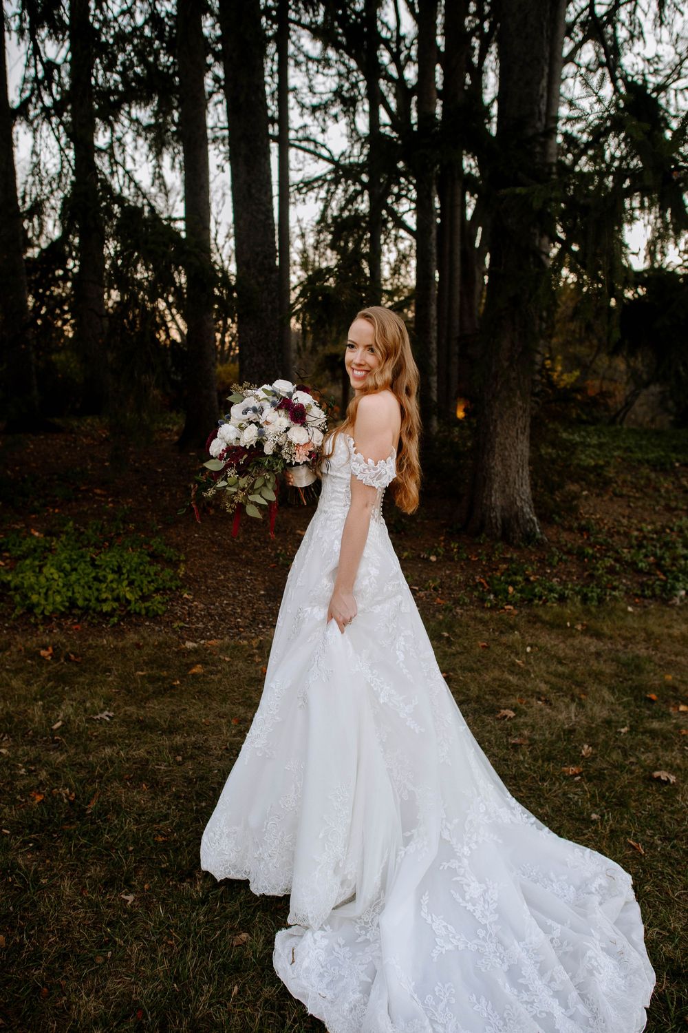 Smiling bride in off-shoulder lace wedding gown holding floral bouquet outdoors at dusk among tall trees.