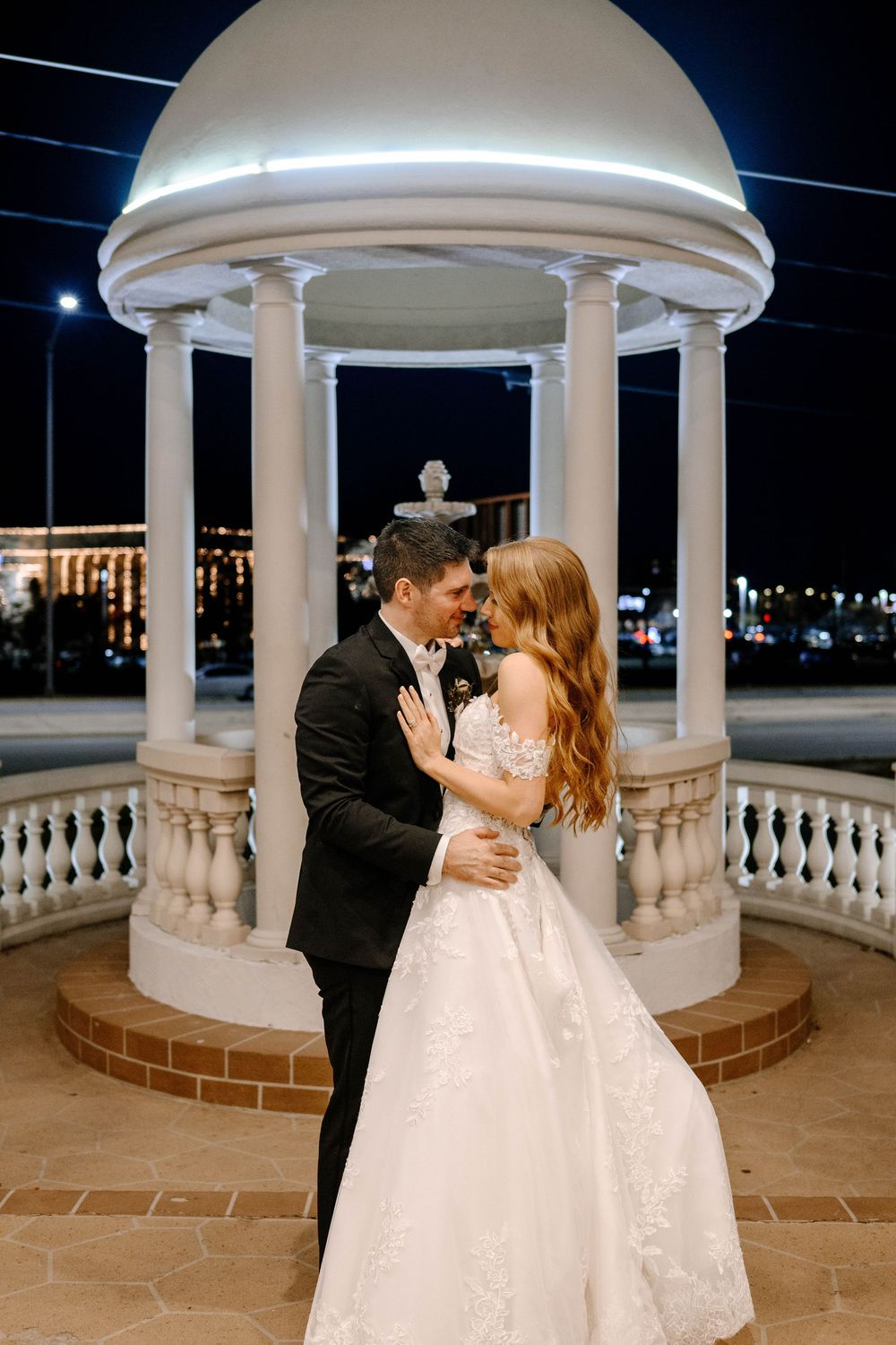 Bride and groom share a romantic moment under a white gazebo at night with city lights in the background.