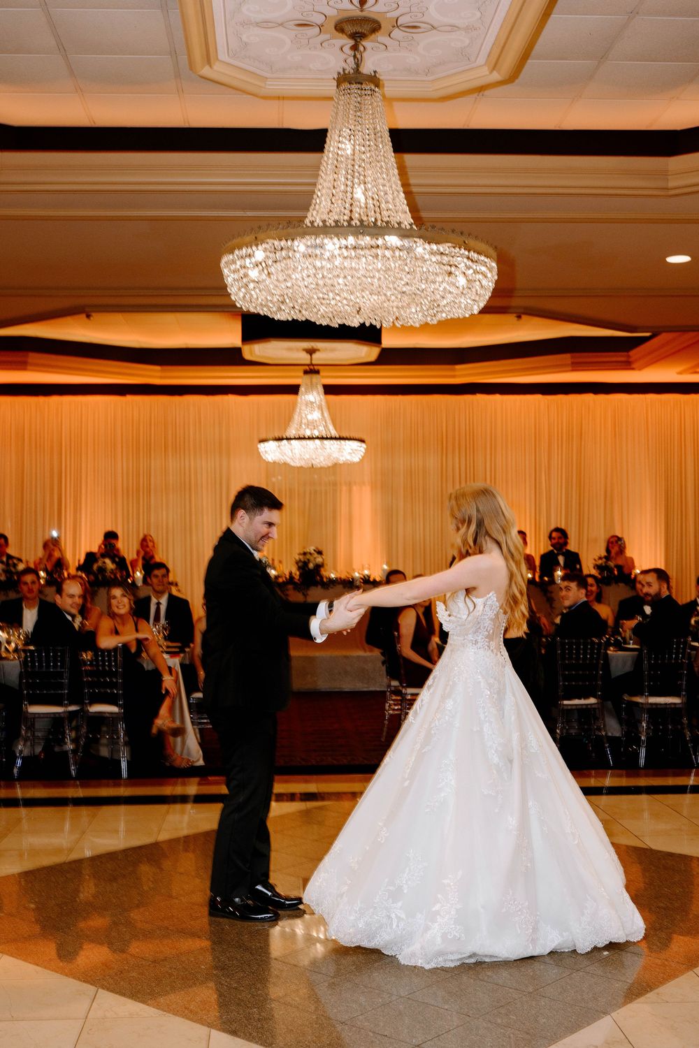 Bride and groom share their first dance under a crystal chandelier in an elegant ballroom wedding reception.