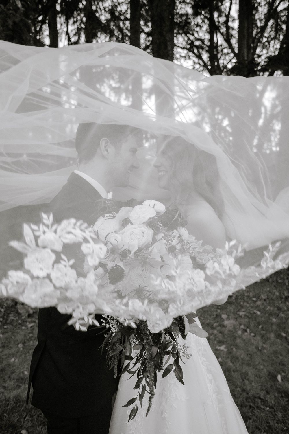 Black and white photo of bride and groom kissing under flowing veil with bridal bouquet.
