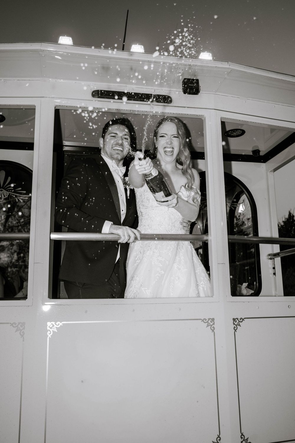 Bride and groom celebrate on a trolley popping champagne in a joyful black and white wedding photo.