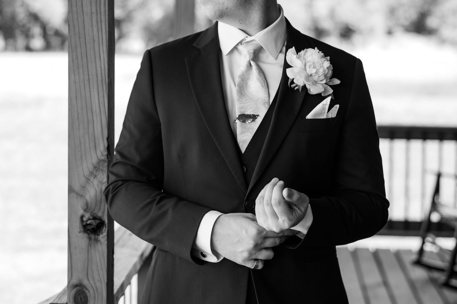 Black and white photo of a groom in a dark suit, silk tie, boutonniere, and pocket square adjusting his cufflinks.