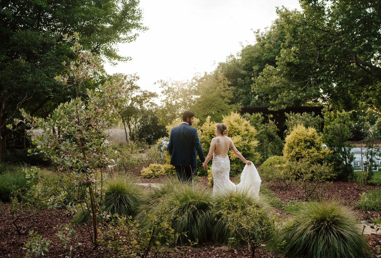 Bride and groom share a romantic moment at golden hour on a gravel path surrounded by lush green trees.