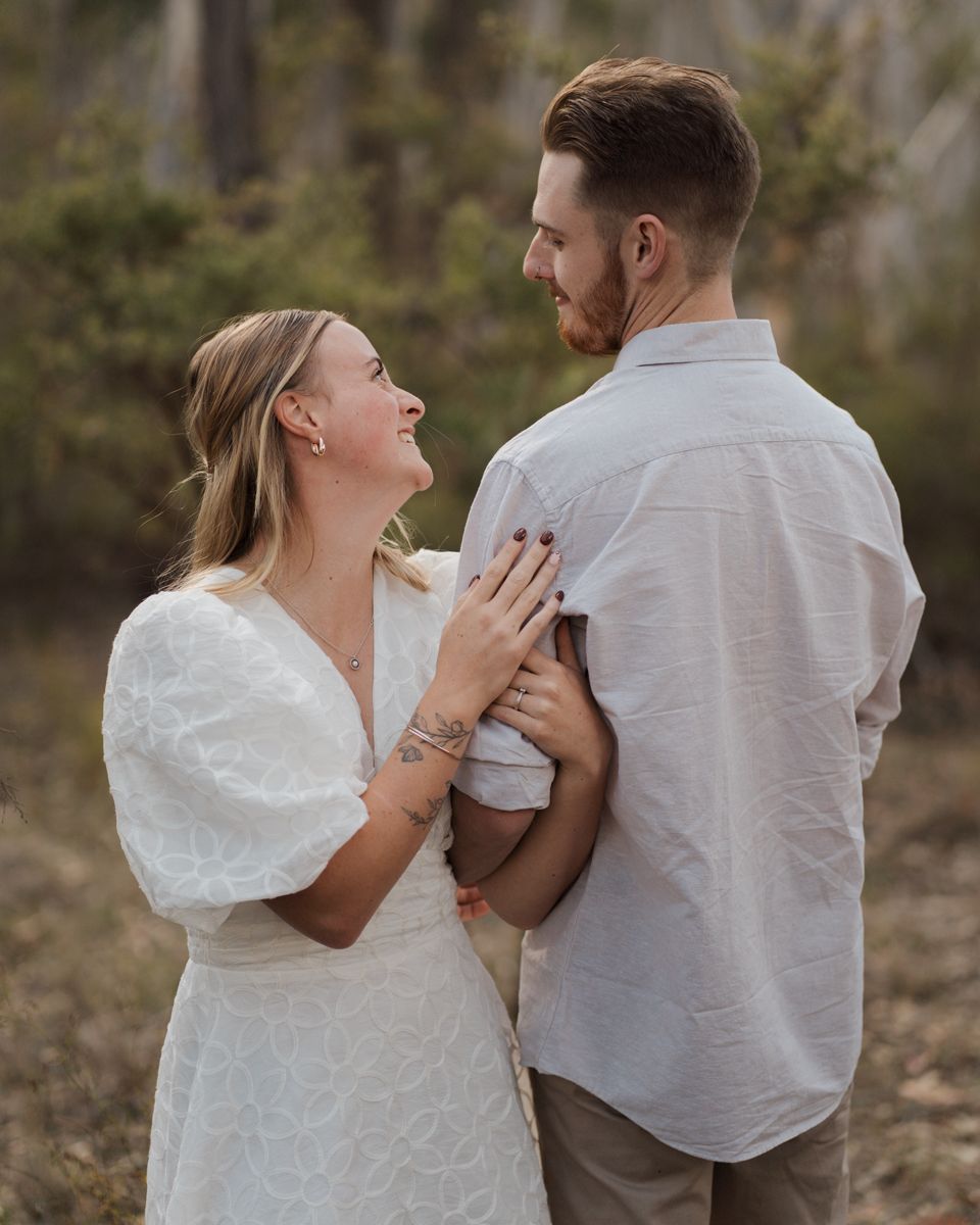 A couple shares a joyful moment outdoors, woman in white dress laughing up at bearded man in light shirt engagement photoshoot rouse hill