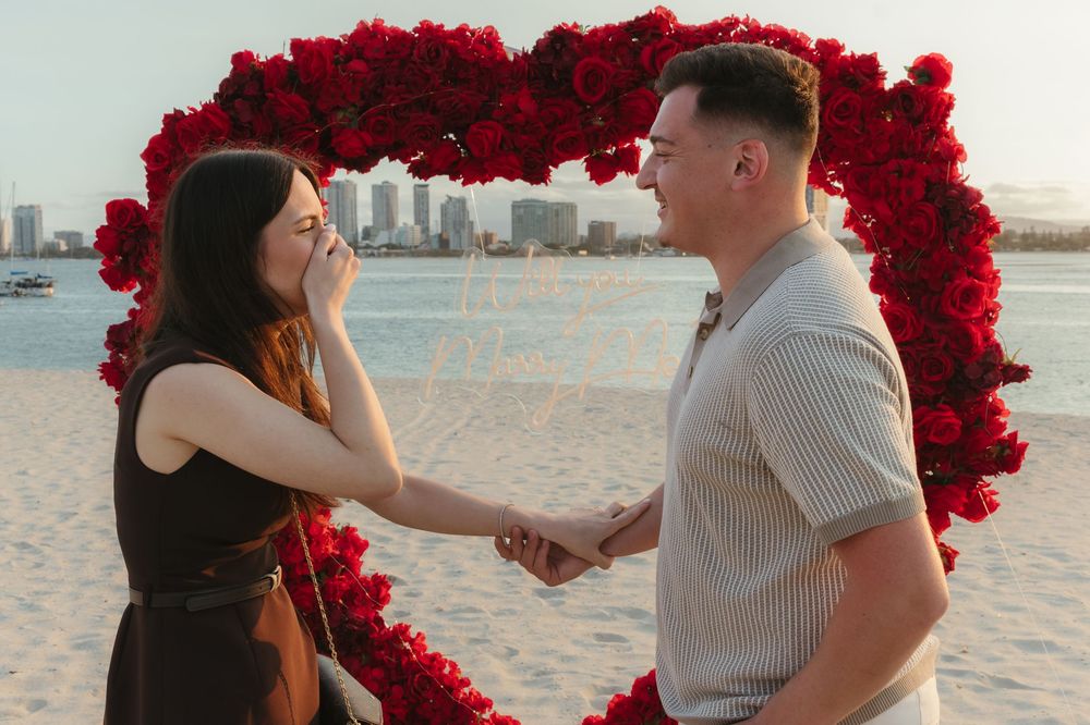 Couple holding hands during beach proposal under red rose heart arch with city skyline backdrop.