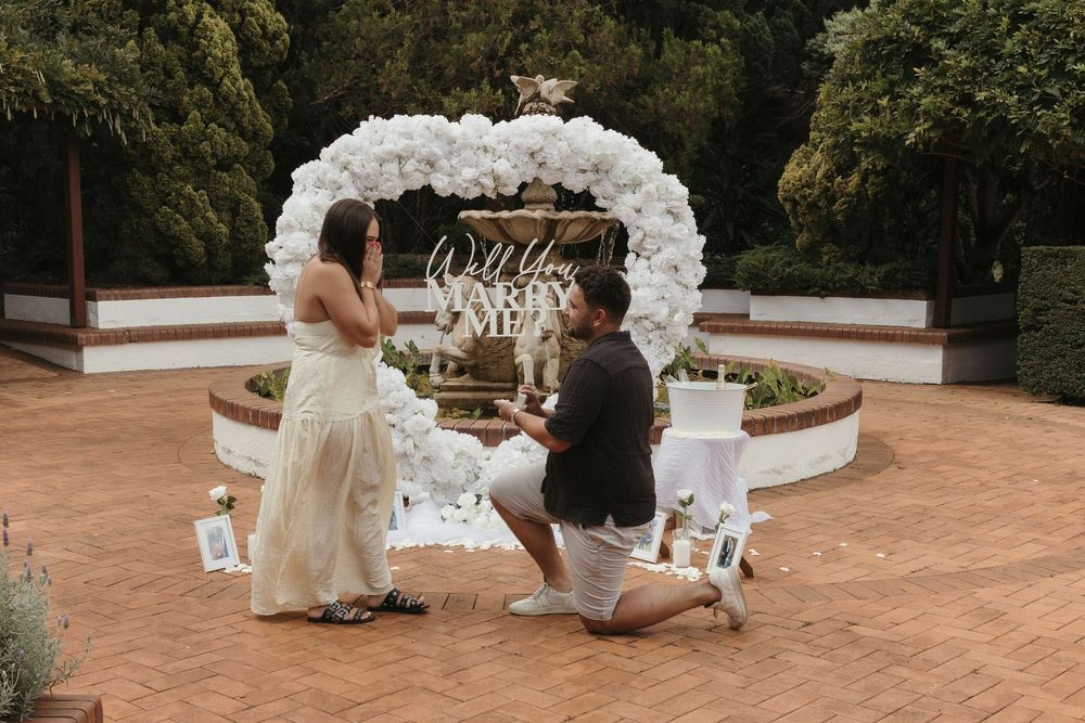 Man proposing to woman on one knee in front of a white balloon arch and fountain in a garden setting.