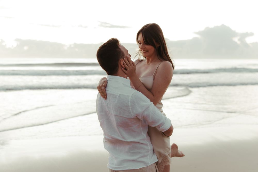 Romantic couple embracing on a beach at sunset, man lifting woman as waves crash behind them.