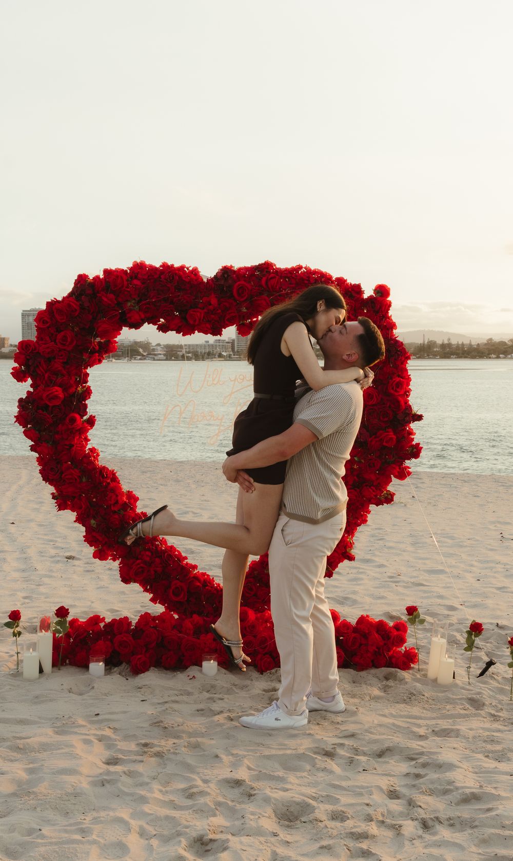 Couple kissing in front of a large red rose heart arch on a beach with candles at sunset.