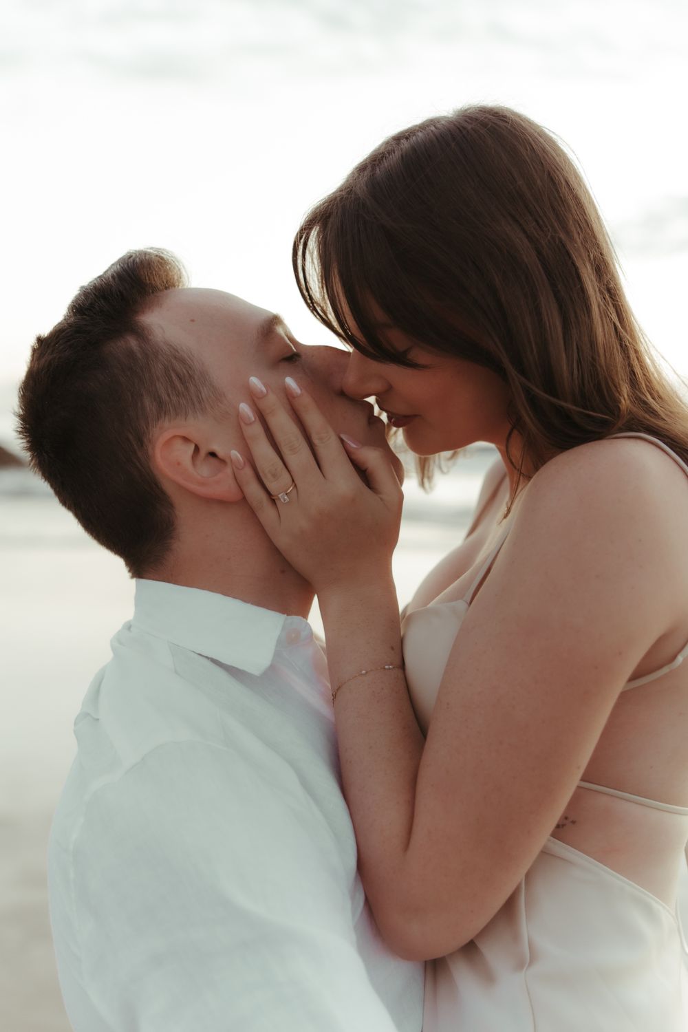 Romantic couple sharing a tender kiss on the beach, woman in pink dress cradling man's face near ocean waves.