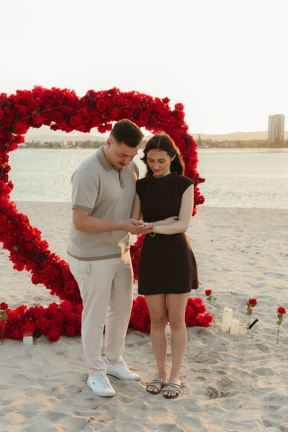Couple sharing a romantic beach proposal moment in front of a large red rose heart arch at sunset.