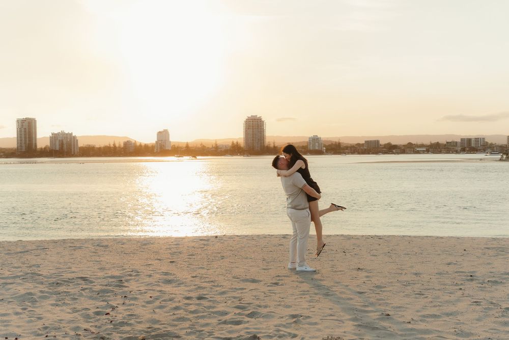 Couple embracing on a sandy beach at golden sunset with city skyline reflecting on calm water behind them.