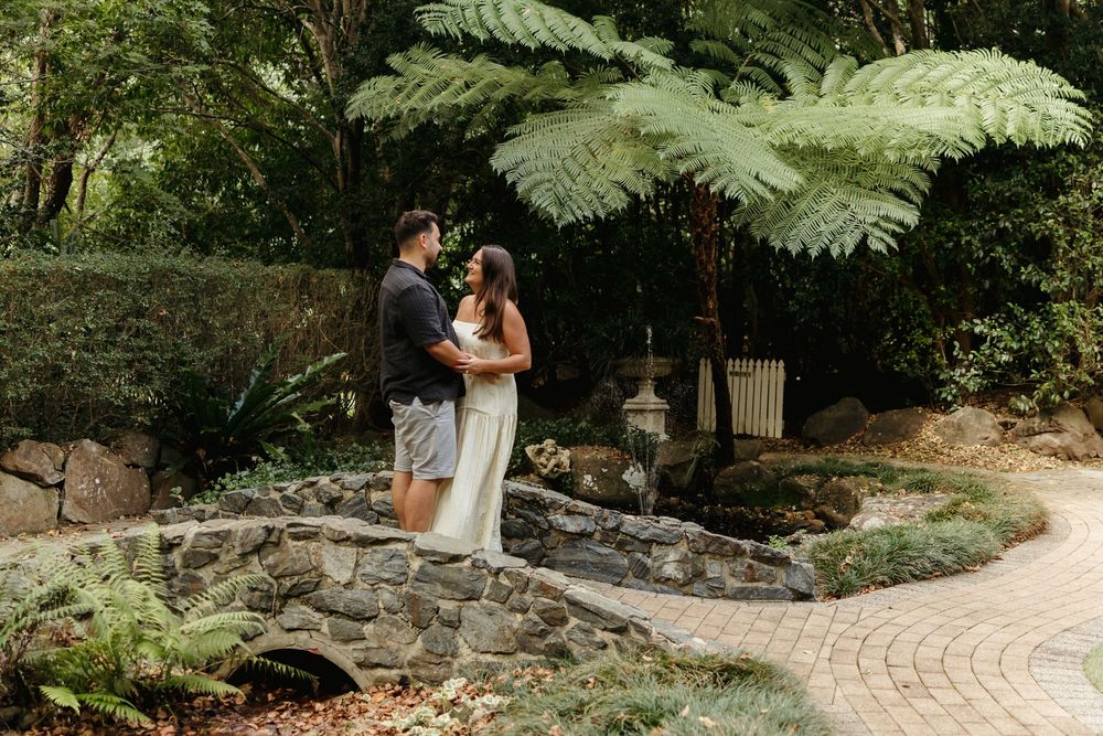 Couple shares a romantic moment in a lush garden with giant fern trees and stone pathway.
