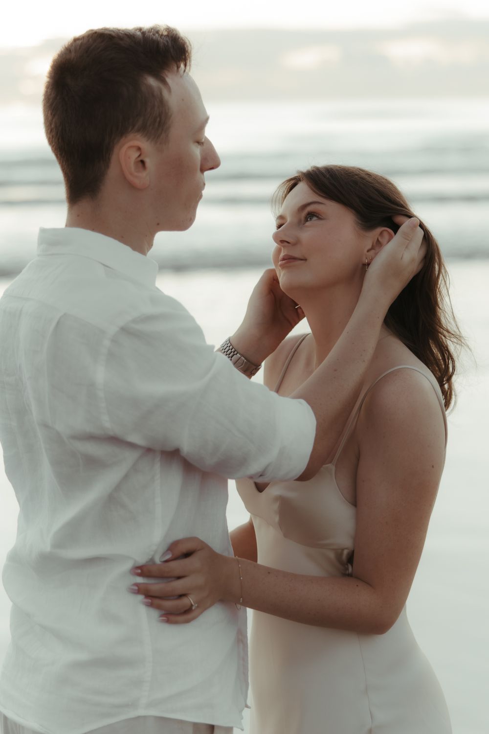 A romantic couple shares a tender moment on a beach, the man gently touching the woman's face as waves crash behind them.