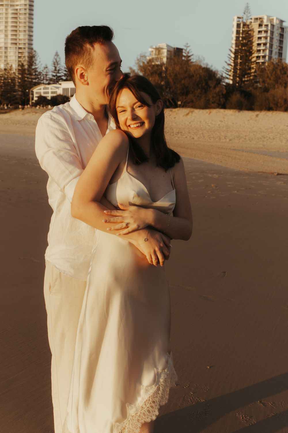 Couple embracing on a sandy beach at golden hour with city buildings in the background.